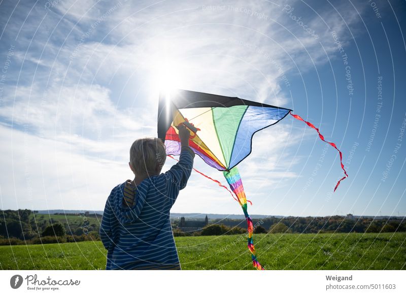 Junge von hinten mit gestreiftem Kapuzenpulli und Blick über Felder und Wiesen lässt einen bunten Drachen steigen Drachensteigen junge Herbst Regenbogen