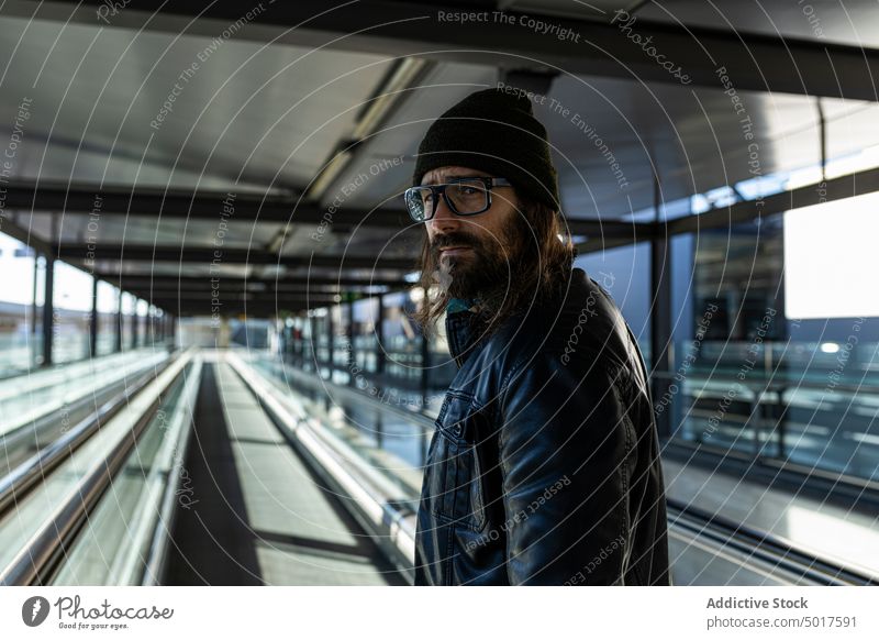 Hipster männlicher Reisender auf dem Laufband im Flughafen Mann reisen Passagier Koffer trendy Stil Erwachsener modern Gepäck Vollbart Hut Lederjacke Ausflug