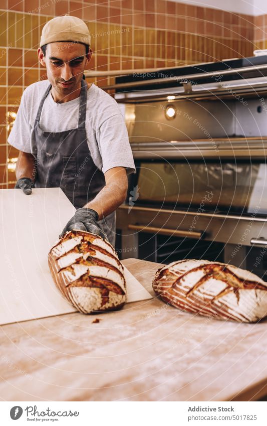 Lächelnder männlicher Koch mit Brot in einer Bäckerei Mann hölzern Tisch gebacken frisch geschmackvoll Ofen heiter Brotlaib Lebensmittel Gebäck lecker Schürze