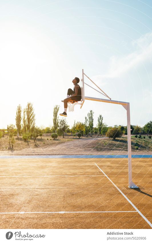 Schwarzer Spieler sitzt auf Basketballkorb und hält Bein Sportler Reifen Spielplatz professionell Sportbekleidung Spielen Athlet Meisterschaft Fähigkeit