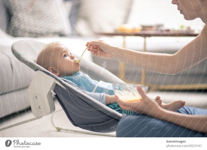 Mutter Löffel füttern ihr Baby Junge Kleinkind Kind in Babystuhl mit Fruchtpüree. Baby feste Nahrung Einführung Konzept. Gesundheit Ernährung Essen Lebensmittel