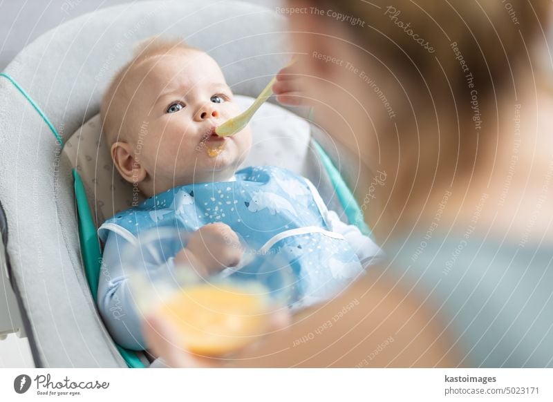 Mutter Löffel füttern ihr Baby Junge Kleinkind Kind in Babystuhl mit Fruchtpüree. Baby feste Nahrung Einführung Konzept. Gesundheit Ernährung Essen Lebensmittel