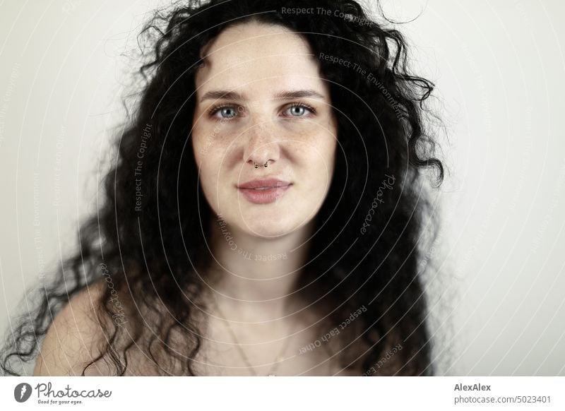 Nahes Portrait einer jungen, dunkelhaarige Frau mit Locken, hellblauen Augen und Sommersprossen vor einer weißen Wand junge Frau brünett lange Haare blass schön