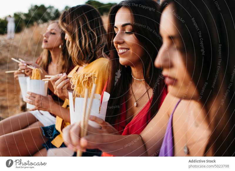 Fröhliche Frauen, die in der Natur asiatisch zu Mittag essen Freundin Mittagessen Spaß Kasten Nudel Asiatische Küche Mahlzeit Essstäbchen Lebensmittel Stein