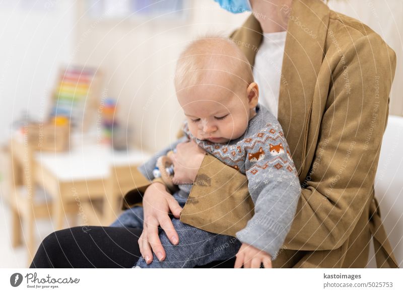 Mutter mit Kleinkind auf dem Schoß, sitzend und wartend vor einer Arztpraxis für eine pädiatrische Untersuchung. Gesundheitskonzept für das Kind Säugling