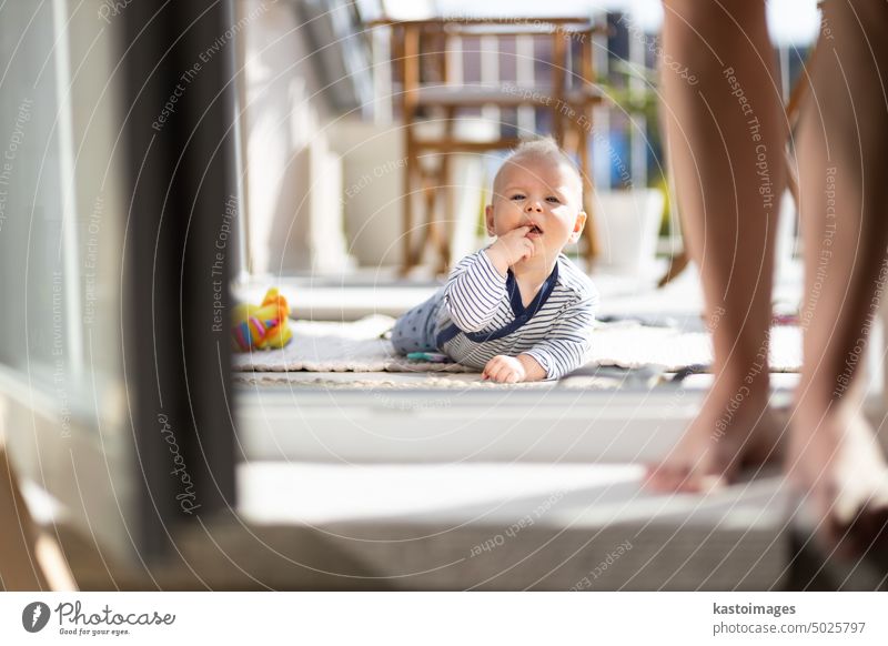 Niedliches kleines Baby, das mit Spielzeug im Freien auf der Terrasse im Sommer spielt und von seiner Mutter im Hintergrund beaufsichtigt wird. Säugling