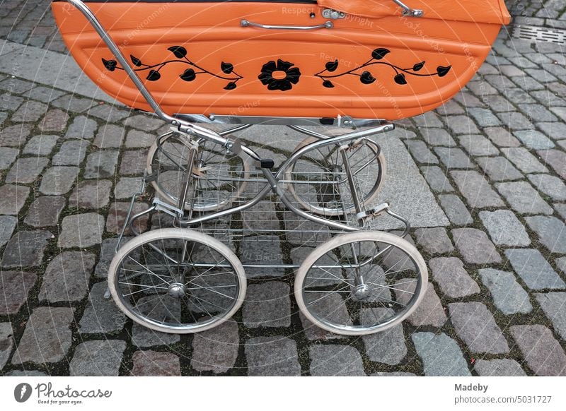 Alter Kinderwagen der Fünfzigerjahre in Orange mit Blumenmuster auf altem grauen Kopfsteinpflaster am Oberstadtmarkt in Marburg an der Lahn in Hessen Wagen