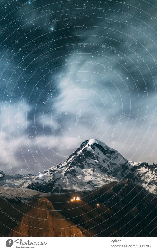 Stepantsminda, Georgien. Real Night Starry Sky With Glowing Stars Over Peak Of Mount Kazbek mit Schnee bedeckt. Berühmte Gergeti Kirche in der Nacht Blitz. Schöne georgische Landschaft im Spätherbst