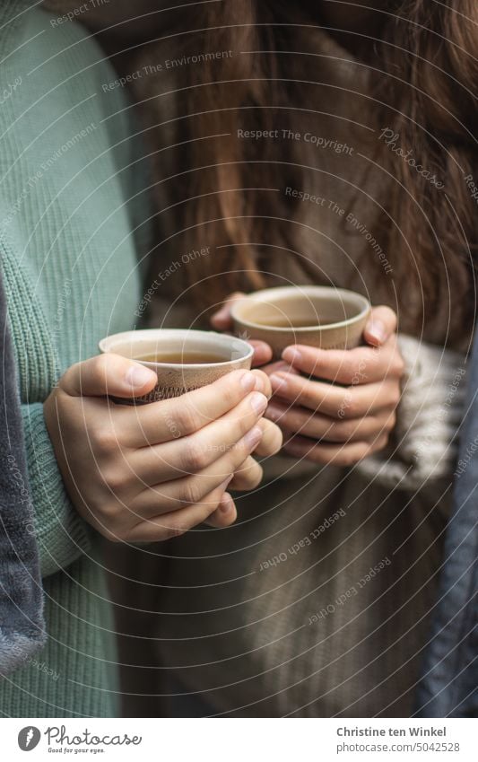 Detailaufnahme zweier junger Frauen in kuscheligen Pullovern mit Teebechern in den Händen festhalten wärmend heißer Tee Heißgetränk gemeinsam aufwärmen Kälte