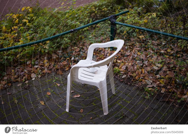 Plastikstuhl mit Tasse steht in einem Winkel grüner Geländerstangen als Sitzplatz auf Steinboden Stuhl Kunststoff Monobloc weiß stapelbar Massenware sitzen