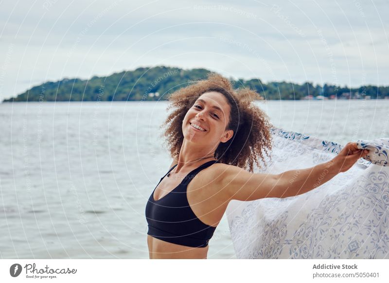 Zufriedene Frau mit wehendem Schal am Meeresufer Glück Wind Freiheit Strand Küstenlinie heiter Sommer positiv Strandbekleidung winken Stoff hispanisch