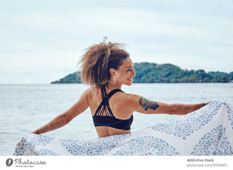 Zufriedene Frau mit wehendem Schal am Meeresufer Glück Wind Freiheit Strand Küstenlinie heiter Sommer positiv Strandbekleidung Lächeln winken Stoff hispanisch