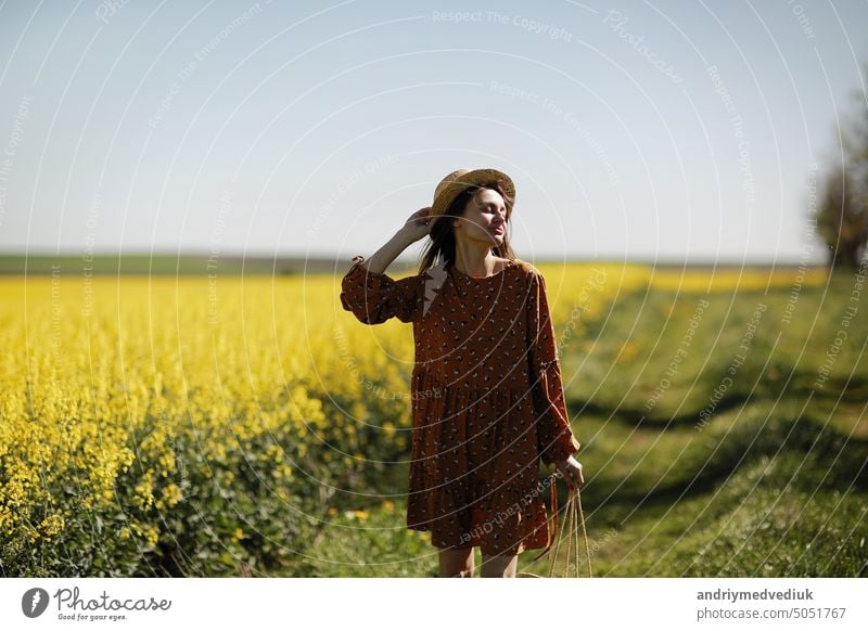 schöne junge Frau geht in einem Feld von gelben Raps. Mädchen brunette lange Haare fliegen im Wind in Kleid und Strohhut gekleidet. Sommerurlaub Konzept Natur