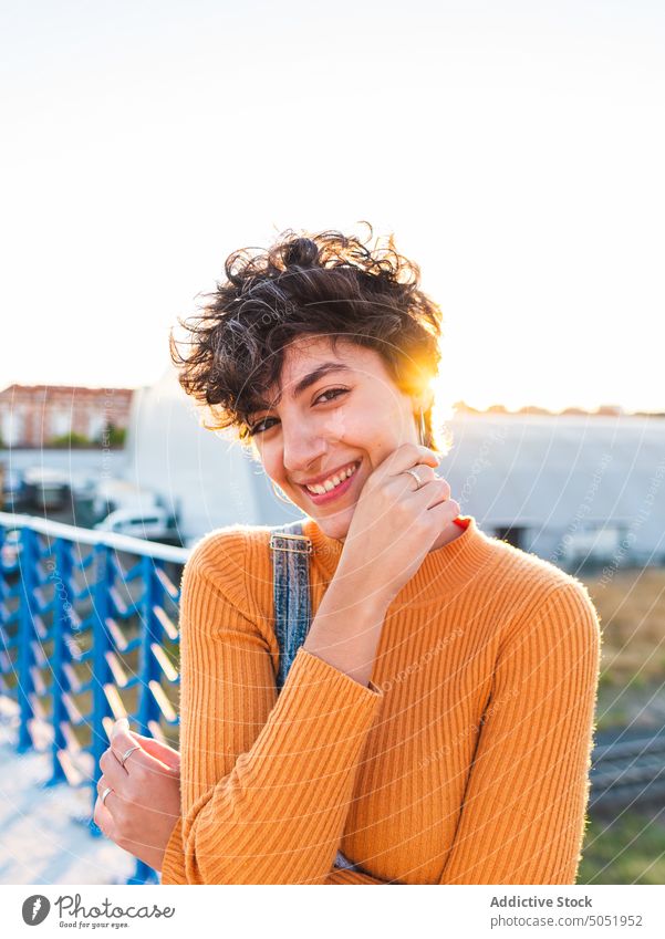 Fröhliche Frau mit kurzen Haaren steht auf einem Steg Lächeln positiv Brücke Zaun Gesicht berühren Glück Sonne Scheune heiter jung Pullover stehen lässig