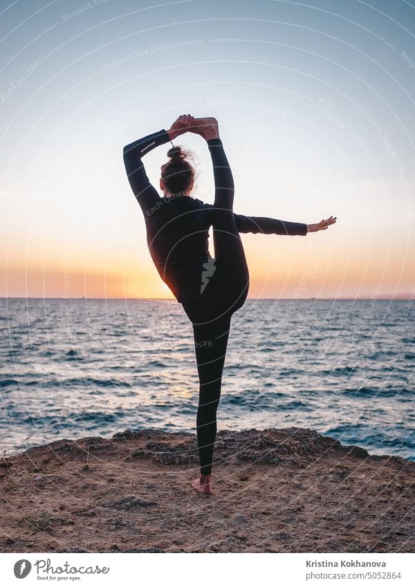 Schlanke Frau in schwarzem Bodysuit übt Yoga in der Nähe von Meer oder Ozean bei Sonnenaufgang Licht. Flexibilität, Stretching, Fitness, gesunder Lebensstil.