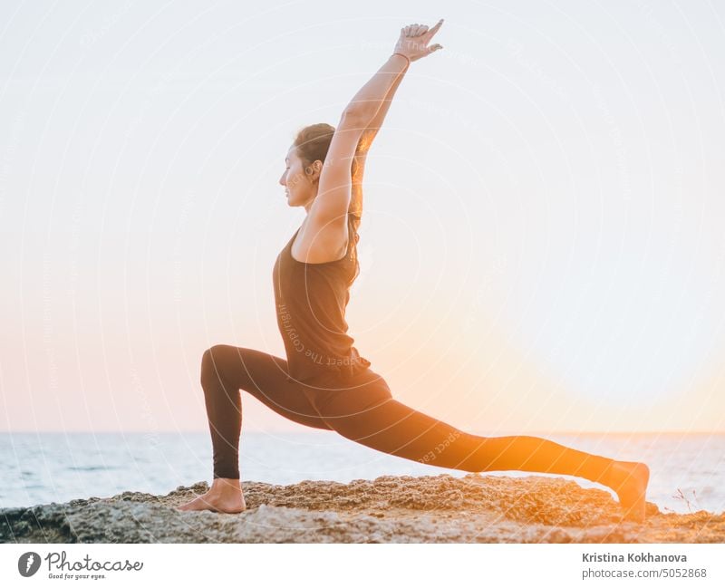 Schlanke Frau in schwarzem Bodysuit übt Yoga in der Nähe von Meer oder Ozean bei Sonnenaufgang Licht. Flexibilität, Stretching, Fitness, gesunder Lebensstil.