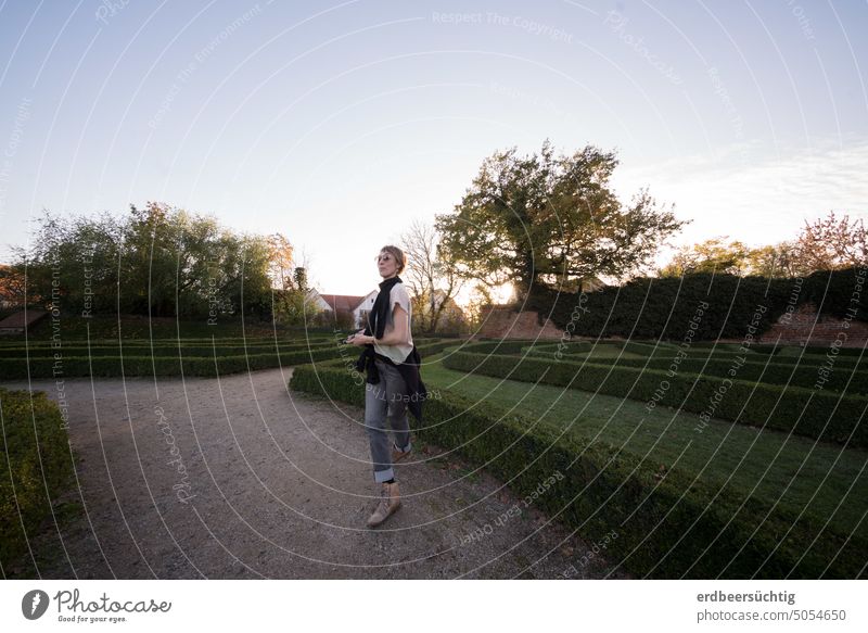 Frau (lust-)wandelt im abendlichen Irrgarten Labyrinth Park Büsche Grün Gartenanlage Weg laufen wandeln Abendlicht Bäume dynamisch tänzerisch Sonnenbrille irren