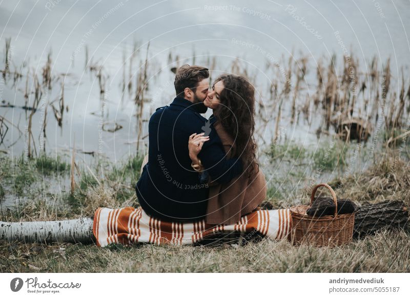 zwei kaukasische Liebhaber sitzen auf Decke am See. Junges Paar umarmt sich am Herbsttag im Freien. Ein bärtiger Mann und lockige Frau in der Liebe. Valentinstag. Konzept der Liebe und Familie. Rückansicht