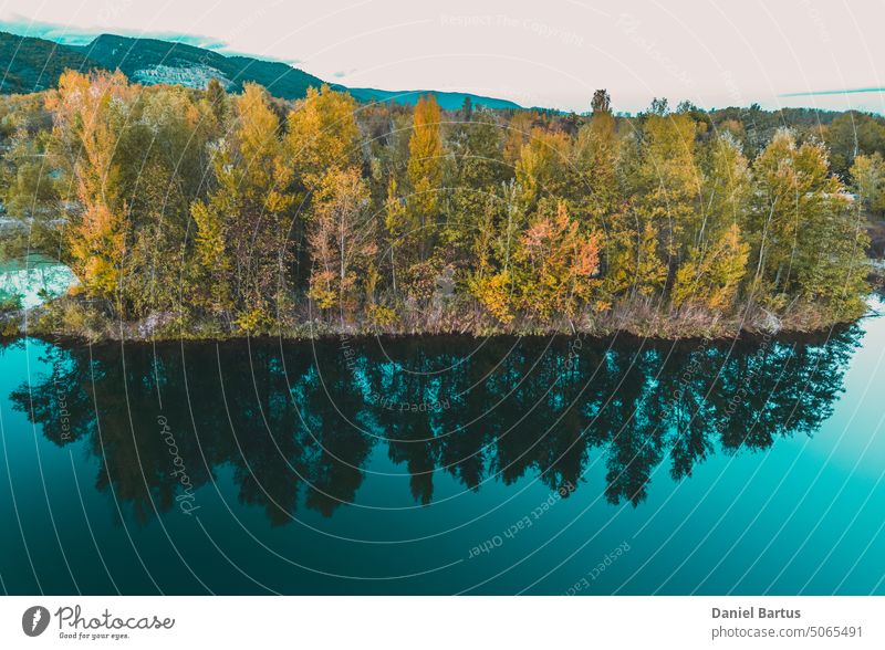 Die Reflexion der Bäume im Herbst auf der Wasseroberfläche, bunte gelb-orange Blätter. Der Wald spiegelt sich in der Wasseroberfläche. Blau-türkisfarbenes Seewasser.