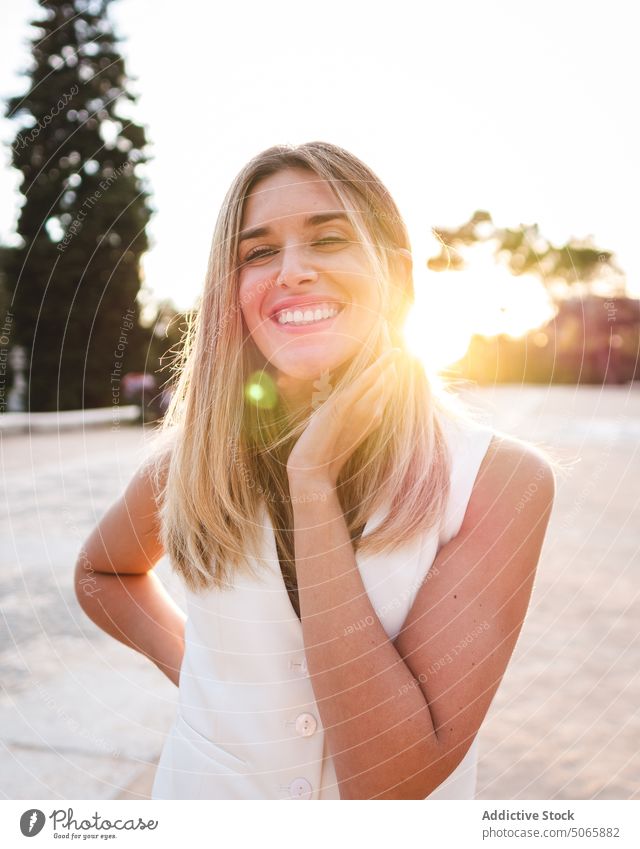 Optimistische Frau berührt Hals bei Sonnenuntergang Park Lächeln Wochenende Sommer Glück Abend Tasthals Vorschein jung Sonnenschein blond heiter Abenddämmerung