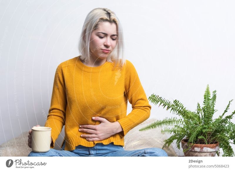 Blonde Frau sitzt auf dem Boden und hält eine Tasse Tee und eine Hand auf dem Bauch blond Übelkeit Sitzen Beteiligung Kaffee Person im Innenbereich Schmerz