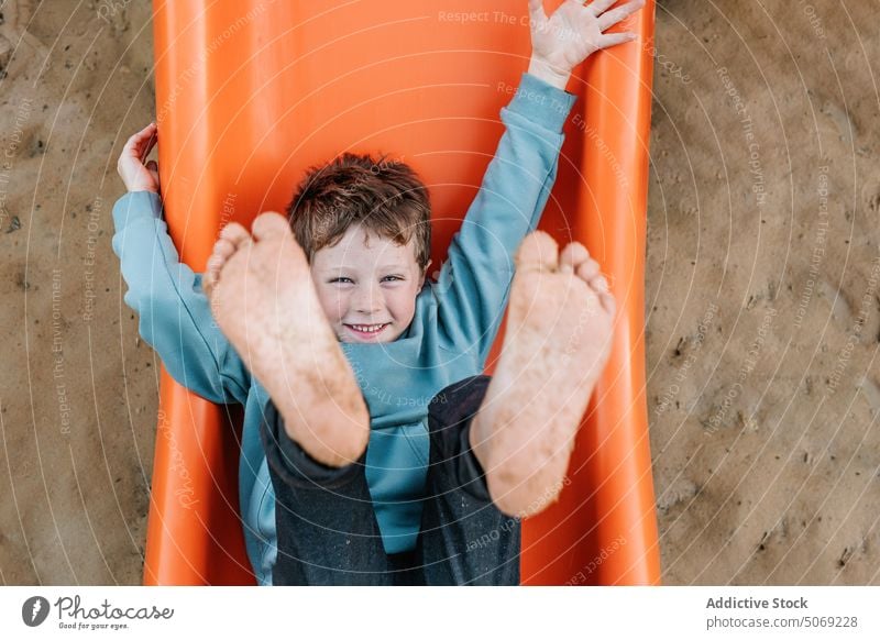 Fröhlicher Junge spielt auf orangefarbener Rutsche spielen Sliden Spielplatz Sand Barfuß zeigen dreckig Glück Wochenende Sommer Kind Boden Zeitvertreib