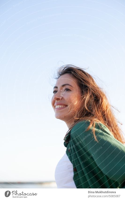 Schöne junge Frau lächelnd mit blauem Himmel auf dem Hintergrund. Kaukasische Frau mit braunen Haaren lächelnd und Blick nach vorne trägt grünes Outfit. Lifestyle candid weibliches Porträt.