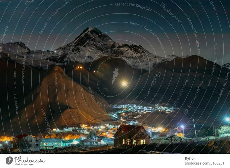 Stepantsminda, Georgien. Peak of Mount Kazbek mit Schnee bedeckt, berühmte Gergeti Kirche und Landschaft Häuser in der Nacht Blitz Ansicht Dorf Stadt malerisch