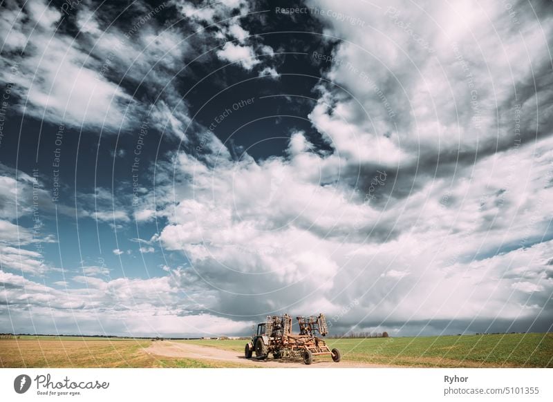 Traktor mit angebauter Egge im sonnigen Frühlingstag. Countryside Rural Road Through Field Meadow Landscape Under Scenic Dramatic Sky With Clouds Before Rain