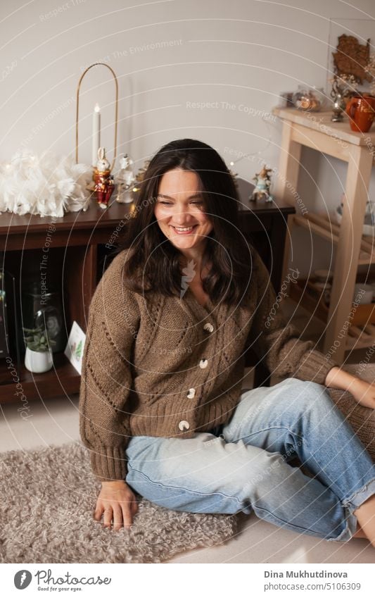 Candid Foto von einer Frau Jeans und Strickjacke sitzen zu Hause für die Weihnachtsferien dekoriert, feiern. Lächelnde Frau mit braunem Haar, lachend. heiter