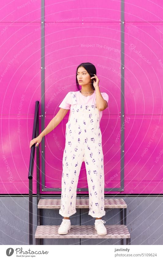 Chinesische Frau mit leerem Blick und ernstem Gesichtsausdruck, die an einer rosa Wand eines modernen Gebäudes steht. asiatisch modisch urban Japanisch Mode