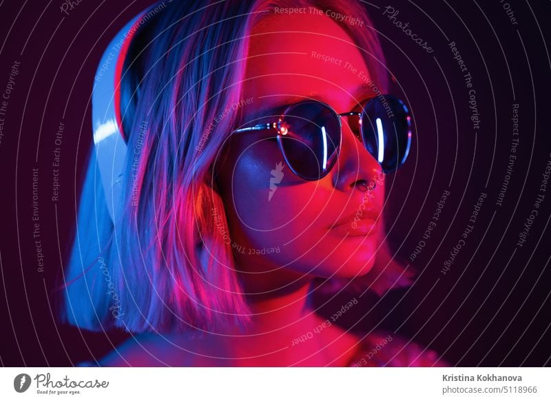 Gorgeous Dame mit gefärbten Haaren und Sonnenbrille hören Musik in Kopfhörer und Gesang mit Neonlicht Hintergrund. Charmante Hipster Mädchen tanzen mit geschlossenen Augen.