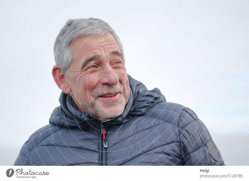 Porträt eines Senioren mit kurzen grauen Haaren, Dreitagebart und verschmitztem Lächeln Mensch Mann Alter Jacke Winter schönes Wetter Himmel Männlicher Senior