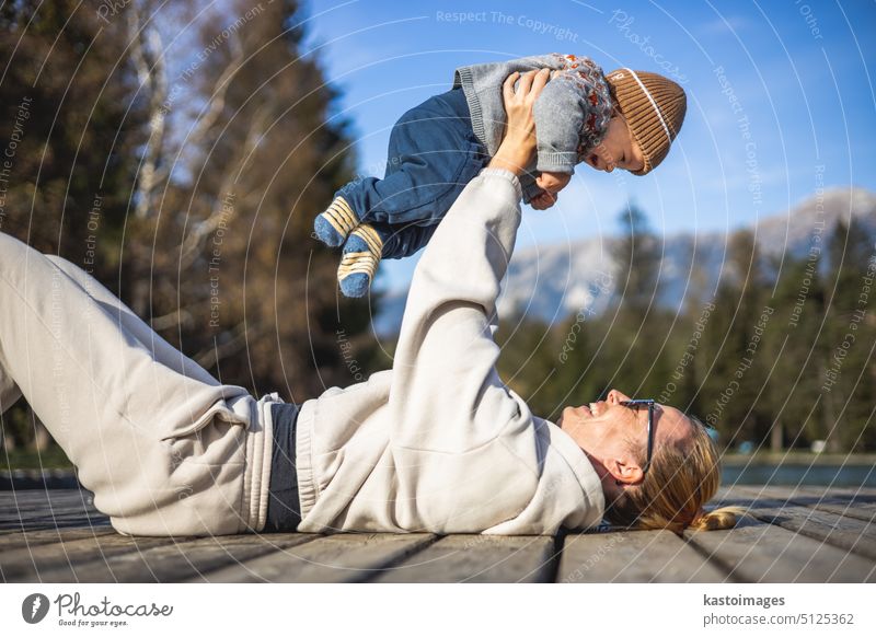 Glückliche Familie. Junge Mutter spielt mit ihrem kleinen Jungen Kleinkind im Freien auf sonnigen Herbsttag. Porträt von Mutter und kleinem Sohn auf Holzplattform am See. Positive menschliche Emotionen, Gefühle, Freude.