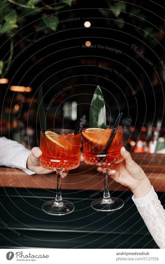 Hände von Frau und Mann klirren, Prost mit Gläsern von Spritz Cocktail. Paar feiert Hochzeit, Jahrestag mit Aperol Spritz Cocktails, mit Orange und Grüns Erfrischendes alkoholisches Getränk