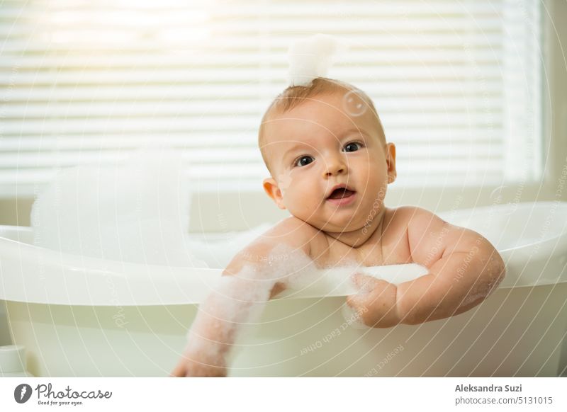 Nettes kleines Baby sitzt in der weißen Badewanne mit Schaum und Seifenblasen. Nimmt ein Bad und spielt mit Spielzeug. Baby-Hygiene. 6 Monate 6 Monate Baby