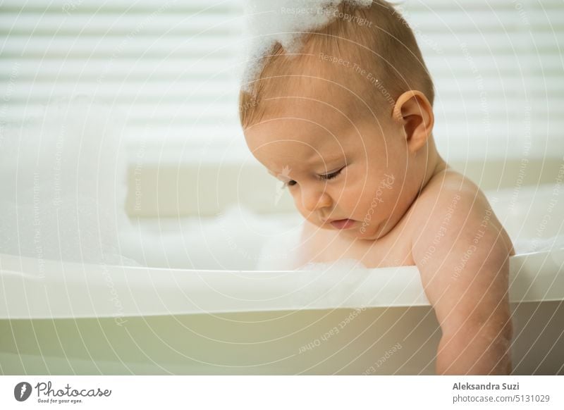 Nettes kleines Baby sitzt in der weißen Badewanne mit Schaum und Seifenblasen. Nimmt ein Bad und spielt mit Spielzeug. Baby-Hygiene. 6 Monate 6 Monate Baby