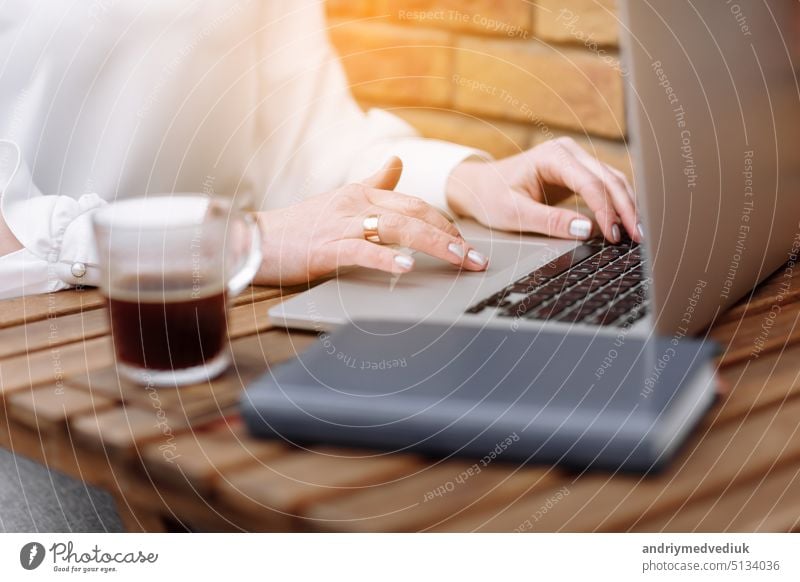 Freelancer Mädchen arbeiten im Café. Cropped Foto der Frau Hände sind mit Laptop im Freien sitzen. Auf dem Tisch Glas Tasse heißen Kaffee und Papier-Notebook. Kaffeepause. Online-Job oder Studium