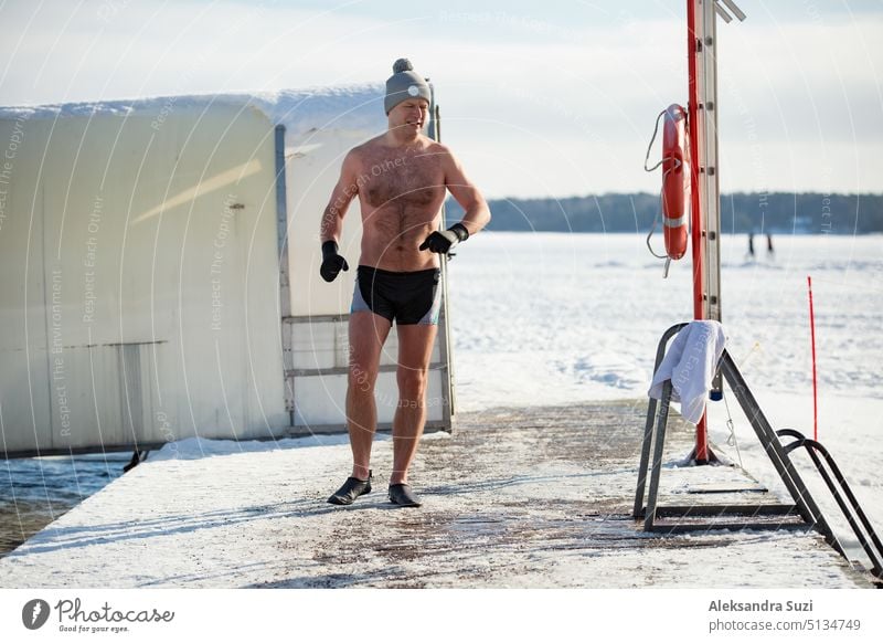 Ein Mann mit warmer Mütze und Handschuhen schwimmt in einem Eisloch und geht am Pier entlang. Winteraktivitäten in Finnland. Gesunder Lebensstil Aktivität