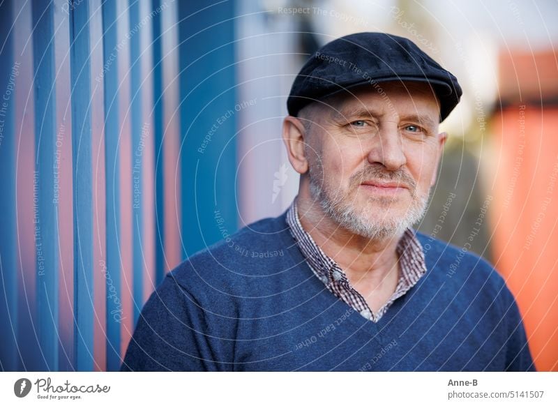 Portrait eines freundlich blickenden Mannes mit blauen Augen, blauem Pullover und blauer Schiebermütze , Bart und direktem Blickkontakt. Der Hintergrund ist eine blaue Holzwand.