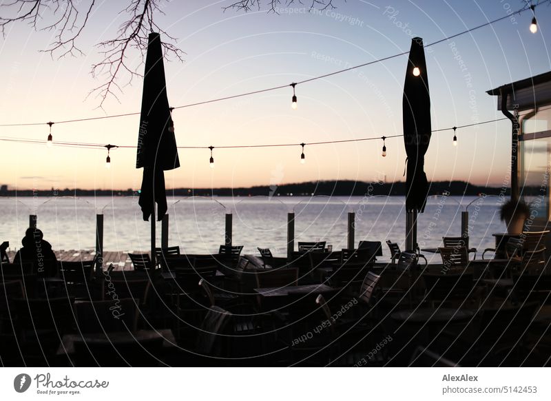 Aussenbereich eines Cafés/Restaurants im Gegenlicht in der Dämmerung am Meer/Ostsee bei Kiel mit Lichterketten und Sonnenschirmen aussen Außenbereich Stühle