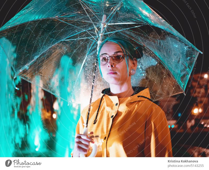 Junges hübsches Mädchen mit blau gefärbtem Haar in gelbem Regenmantel und mit transparentem Regenschirm steht in der Nähe von Brunnen. Nacht Neon-Beleuchtung der Stadt. Porträt von stilvollen Hipster mit Brille.