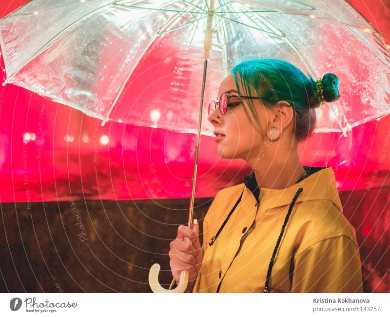 Junges hübsches Mädchen mit blau gefärbtem Haar in gelbem Regenmantel und mit transparentem Regenschirm steht in der Nähe von Brunnen. Nacht Neon-Beleuchtung der Stadt. Porträt von stilvollen Hipster mit Brille.