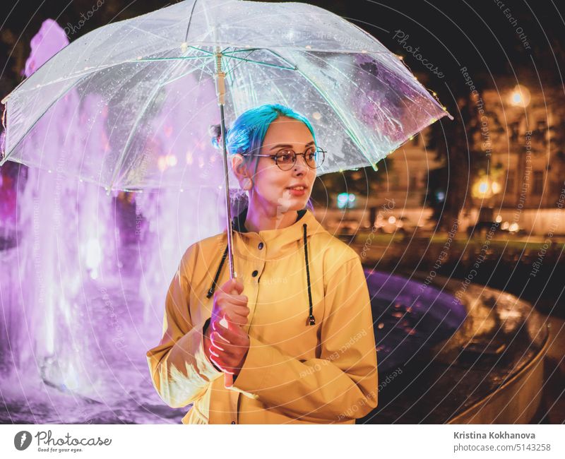 Junges hübsches Mädchen mit blau gefärbtem Haar in gelbem Regenmantel und mit transparentem Regenschirm steht in der Nähe von Brunnen. Nacht Neon-Beleuchtung der Stadt. Porträt von stilvollen Hipster mit Brille.