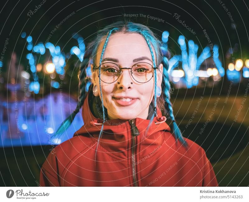 Junge schöne Mädchen mit blau gefärbten ungewöhnliche Haare, transparente Brille, Piercing stehen in der Nacht Straße mit Neonröhren Hintergrund. Porträt des glücklichen niedlichen stilvollen Teenager.