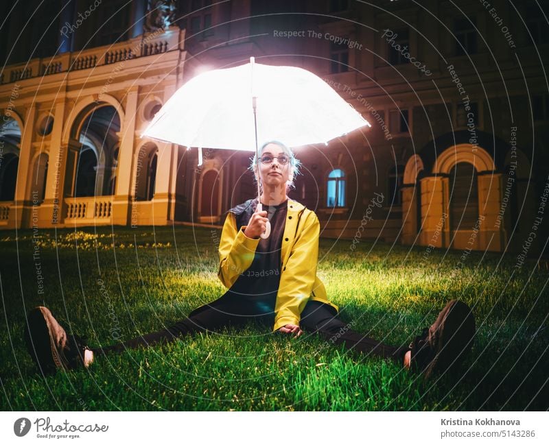 Junges hübsches Mädchen mit blau gefärbten Haaren in gelbem Regenmantel und mit transparentem leuchtendem Regenschirm sitzt auf grünem Gras in der Nähe eines historischen Gebäudes. Porträt von stilvollen Hipster mit Brille.