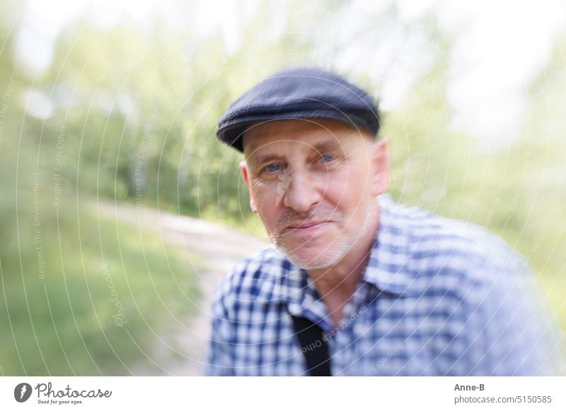 Mittfünfziger mit blauen Augen, blaukariertem Hemd und einer blauen Schiebermütze im Grünen schaut mich verschmitzt an .Lensbaby mit schön viel Unschärfe im Bild.