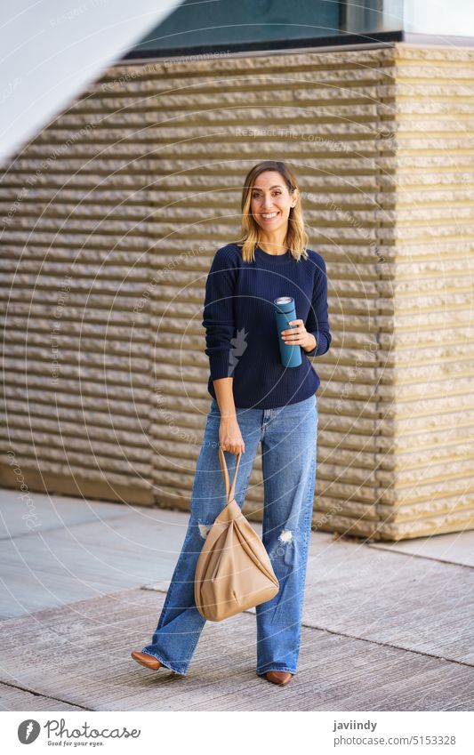 Fröhliche Frau gegen modernes Gebäude Straße Stil Lächeln Outfit tagsüber Tasche Thermoskanne Straßenbelag Pullover Jeanshose urban Struktur positiv