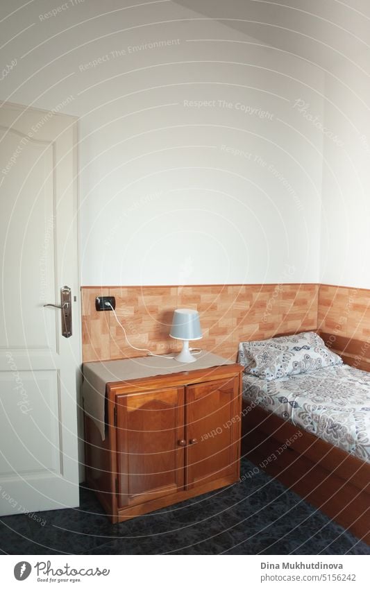 Sehr einfaches Hotelzimmer mit alten Holzmöbeln und weißen Wänden. Interieur eines billigen Zimmers zur Miete in einem Haus. Vermietung von Immobilien und Wohnungen. Kleines Hotel oder Gästehaus Interieur Zimmer vertikale Ansicht mit Einzelbett und Nachttisch mit Lampe.