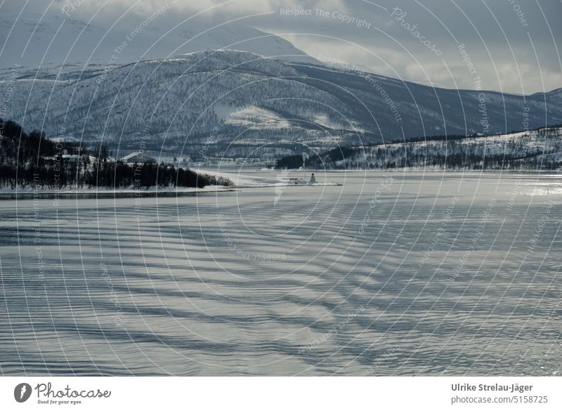 Hafeneinfahrt mit kleinem Leuchtturm und Siedlung an der norwegischen Küste Winter winterliche Küste kaltes Meer Wellen auf dem Meer graue Wolken